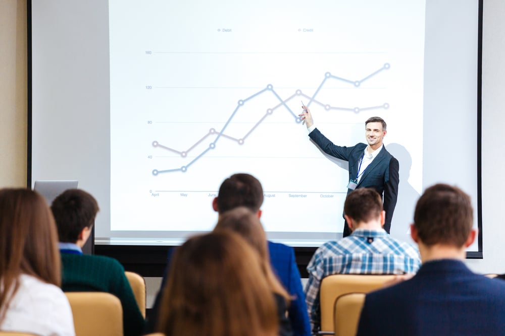 Smiling handsome speaker standing and explaining graphs on business conference in meeting hall-1